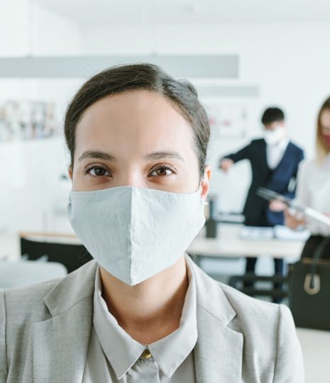 Young mixed-race businesswoman in protective mask looking at you on background of her colleagues by their desks going to work